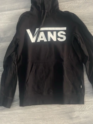 Svart hoodie från Vans med logga - Svart hoodie från Vans med stor vit logga över bröstet. Klassisk känguruficka framtill och justerbar huva med snören. Tillverkad i mjukt material som passar perfekt för en avslappnad streetwear-look.