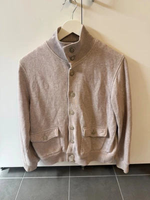 Gran Sasso Cardigan, Stl 46/S - Säljer en stilren beige kofta med ståkrage och stora knappar framtill. Koftan har två fickor med lock och knapp, samt ribbade muddar vid ärmslut och nederkant. Perfekt för lager på lager och enkel att matcha med olika outfits.
