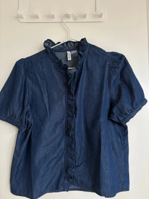 Mörkblå blus med volangdetaljer - Snygg mörkblå blus i denim-look med korta puffärmar och volang längs krage och knappslå. Blusen har en avslappnad passform och är perfekt för dig som gillar en trendig men enkel stil. Tillverkad i bomull för en skön känsla.
