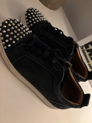 Mörkblå loubs med nitar och röd sula - Säljer ett par svarta sneakers med coola silverfärgade nitar på tån och klassisk röd sula. Skorna har svarta skosnören och en snygg kontrast mellan mockaliknande material och vita sulor. Perfekta för dig som vill sticka ut med din stil.