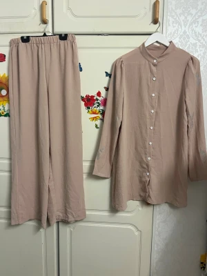 Beige set med långärmad skjorta - Säljer ett beige set med långärmad skjorta och matchande byxor. Skjortan har ståkrage, knappar framtill och puffiga axlar. Byxorna har resår i midjan och är vida i modellen. Materialet ser ut att vara mjukt och luftigt, perfekt för en avslappnad look.