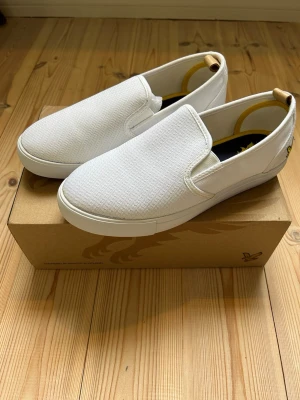 Vita slip-on sneakers från Lyle & Scott - Snygga vita slip-on sneakers från Lyle & Scott med gul broderad logga på hälen. Skorna har en enkel och clean design i textilmaterial med elastiska paneler på sidorna för enkel påtagning. Perfekta för en avslappnad och stilren look.