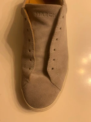Beige mockasneaker från MARCH - Stilren beige sneaker i mocka från MARCH. Skon har en klassisk låg siluett med vit sula och hål för snörning. Perfekt för dig som gillar minimalistisk och clean stil. Ovandelen är i mjukt mockamaterial.