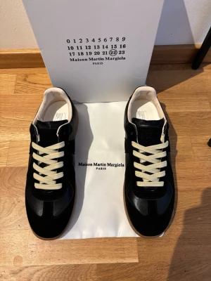 Maison Margiela svarta sneakers - Svarta sneakers från Maison Martin Margiela med vita snören och beige insida. Klassisk låg modell med stilren design och gummisula. Materialet ser ut att vara en mix av läder och mocka för en lyxig känsla.