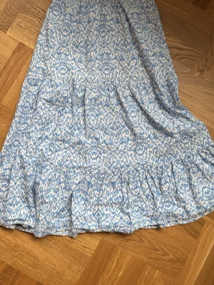 Blåvit mönstrad kjol från Lindex - Säljer en blåvit kjol från Lindex med elastisk midja och ett snyggt abstrakt mönster. Kjolen har volangdetaljer och är gjord i ett lätt och luftigt material, perfekt för varma dagar. Passar dig som gillar en avslappnad men trendig stil.