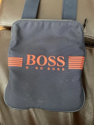 Marinblå axelväska med röd logga - Snygg marinblå axelväska från Hugo Boss med breda axelband och röd logga framtill. Väskan har en enkel, fyrkantig form och är tillverkad i slitstarkt tygmaterial. Perfekt för att bära det viktigaste och ger en clean streetstyle-look.