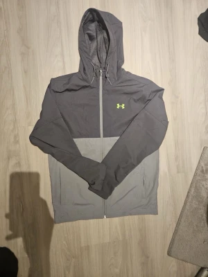 Grå vindjacka från Under Armour - Snygg tvåtonad grå vindjacka från Under Armour med huva och dragkedja framtill. Jackan har meshfoder och en gul logga på bröstet. Perfekt för blåsiga dagar och sportiga outfits.
