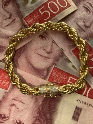Guldpläterat Cordell Armband med Diamantlås  - Säljer ett chunky armband i guldfärgad metall med tvinnad repdesign. Låset är dekorerat med små gnistrande stenar för extra bling. Perfekt för dig som gillar statement-smycken och vill sticka ut lite extra.
