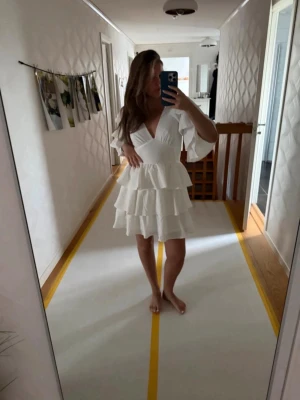 Vit volangklänning med v-ringning - Superfin vit kortklänning med flera volanger och v-ringning både fram och bak. Klänningen har vida trekvartsärmar och en öppen rygg med knappdetalj. Perfekt för sommarens alla tillfällen och ger en romantisk vibe.