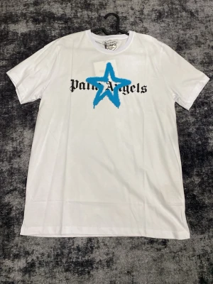 Vit Palm Angels t-shirt med blå stjärna - Säljer en vit t-shirt från Palm Angels med svart logga och en stor blå stjärna tryckt över bröstet. Klassisk rund hals och korta ärmar. Perfekt för dig som gillar streetwear och vill sticka ut med en unik design.