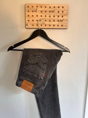 Svarta raka jeans från Levi's - Säljer ett par svarta raka jeans från Levi's med klassisk femficksdesign och läderpatch bak. Jeansen har en diskret tvättad look och är tillverkade i kraftig denim. Perfekta för en avslappnad och stilren outfit.