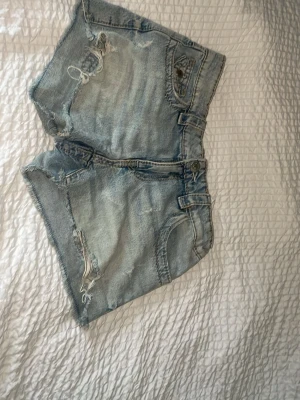 Ljusblå jeansshorts med broderad ficka - Säljer ett par ljusblå jeansshorts med råa kanter och slitningar. Shortsen har en snygg broderad bakficka med paisleymönster och silverdetaljer. Perfekta för sommaren och ger en avslappnad vibe till din outfit.