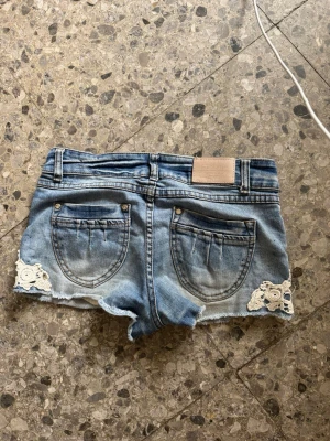 Lågmidjade jeansshorts med spetsdetaljer - Säljer ett par ljusblå denimshorts med låg midja och rå kant. Shortsen har två bakfickor och vita spetsapplikationer på sidorna för en extra söt touch. Perfekta för varma dagar och en avslappnad look.