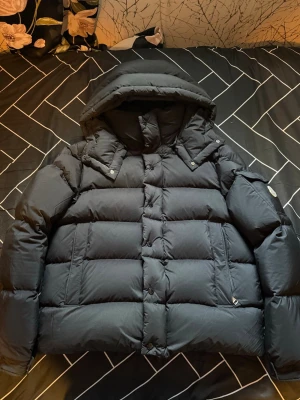 Moncler vezere  - Säljer en svart dunjacka från Moncler med avtagbar huva och klassisk quiltad design. Jackan har dragkedja och tryckknappar framtill, samt fickor med dragkedja. Insidan har glansigt foder och Moncler-logga. Perfekt för kalla vinterdagar.