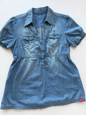 Blå jeansblus från edc - Snygg blå jeansblus från edc med korta ärmar, v-ringning och knappar framtill. Två bröstfickor med lock och en markerad midja ger en avslappnad men trendig look. Tillverkad i mjukt denimtyg som känns bekvämt hela dagen.