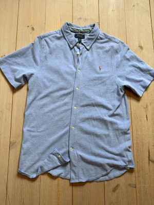 Ljusblå kortärmad skjorta Polo Ralph Lauren - Kortärmad skjorta från Polo Ralph Lauren i ljusblå bomull. Klassisk button-down krage, knappar framtill och liten broderad logga på bröstet. Skjortan är i XL i barnstorlek men sitter som S i vuxenstorlek.