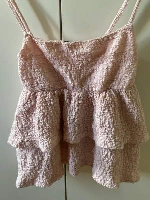 Ljusrosa volangtop från Gina Tricot - Supersöt ljusrosa topp från Gina Tricot med smala axelband och volangdetaljer. Toppen har en skrynklig struktur och är perfekt för varma dagar. Passar dig som gillar en feminin och trendig stil.