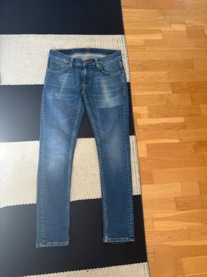 Blå skinny jeans från nudie jeans - Säljer ett par blå skinny jeans från nudie jeans med klassisk femficksmodell och snygga slitningar framtill. Jeansen har normal midja, bälteshällor och dragkedjegylf. Perfekta till en avslappnad och trendig look. I modell tight terry