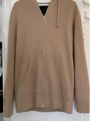 Beige ziphoodie med fickor - En beige hoodie med dragkedja framtill och två stora fickor. Hoodien har huva med dragsko och långa ärmar. Tillverkad i mjukt sweatshirtmaterial.