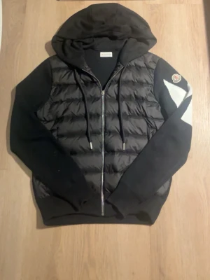 Svart hoodie från Moncler med dunpanel - Svart hoodie från Moncler med quiltad dunpanel framtill, dragkedja, huva med dragsko och Moncler-logga på ärmen. Ärmar och rygg i mjukt sweatshirtmaterial, medan framsidan har en glansig, vadderad look. Snygg detalj med vit kontrast på ena ärmen.