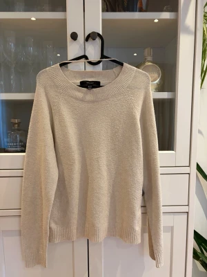 Beige stickad tröja med rund hals - En enkel och stilren beige stickad tröja med rund halsringning och långa ärmar. Tröjan har ribbade muddar vid ärmslut och nederkant, vilket ger en klassisk look. Perfekt att matcha med jeans eller kjol för en avslappnad stil. Nopprig men syns 