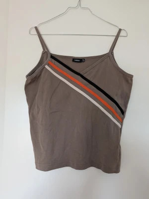 Beige linnetopp med ränder från Lindex - Säljer en beige linnetopp från Lindex med smala axelband och diagonala ränder i svart, orange och vitt framtill. Toppen har en enkel och stilren design, perfekt för varma dagar. Materialet känns mjukt och lätt mot huden.