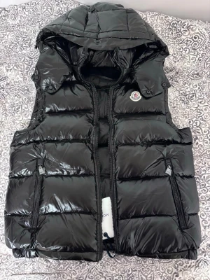 Svart dunväst med huva från Moncler - Snygg svart dunväst från Moncler med glansig finish, avtagbar huva och två dragkedjeförsedda fickor. Klassisk Moncler-logga på bröstet och quiltad design som ger en modern look. Perfekt för lager-på-lager-stil. Pris kan diskuteras 
