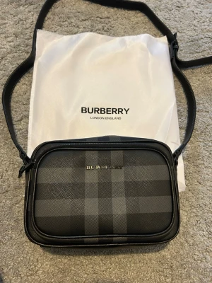Svart  Burberry väska - Snygg svart axelväska från Burberry med klassiskt rutigt mönster i grått och svart. Väskan har justerbar axelrem, två fack med dragkedja och Burberry-logga framtill. Perfekt för dig som vill ha en stilren och tidlös accessoar.
