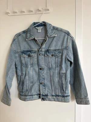 Ljusblå jeansjacka från H&M - Klassisk ljusblå jeansjacka från H&M Adorables med raka sömmar, två bröstfickor med lock och knappar samt två sidofickor. Jackan har en tidlös design med metallknappar framtill och en avslappnad passform. Perfekt för att lyfta vilken outfit som helst.