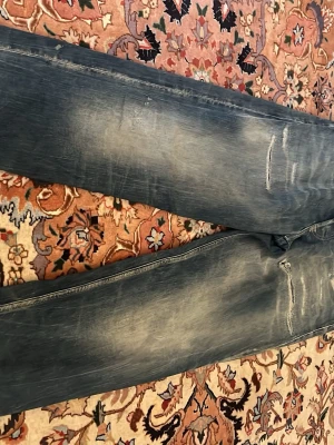 Blå slitna jeans från Diesel - Säljer ett par blå jeans från Diesel med slitningar och tvättade detaljer för en cool, avslappnad look. Klassisk femficksmodell med synliga sömmar och diskreta rivmärken på benen. Perfekta för dig som gillar streetwear och en trendig stil.