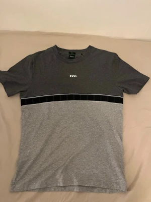 Grå BOSS t-shirt med logga - Snygg t-shirt från BOSS i två nyanser av grått med vit logga på bröstet. T-shirten har en svart rand med diskret mönster över bröstet och rund halsringning. Tillverkad i mjukt bomullsmaterial för en bekväm känsla.