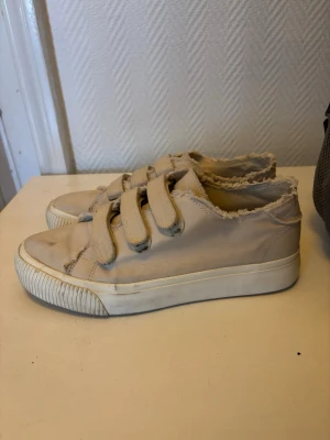 Beige sneakers med kardborreband - Säljer ett par beige sneakers från Even&Odd med tre breda kardborreband och råa, fransiga kanter. Skorna har en tjock vit sula och är tillverkade i tygmaterial. Perfekta för en avslappnad och trendig stil.