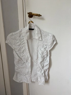 Vit prickig blus från Zara - Söt vit blus från Zara med svarta små prickar och korta puffärmar. Blusen har en klassisk krage och rynkade detaljer framtill som ger en snygg siluett. Perfekt till jeans eller kjol för en trendig look.