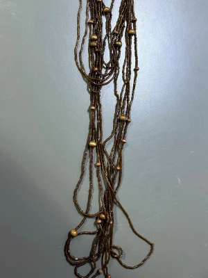 Brunt halsband med pärlor - Säljer ett långt, flersträngat halsband i bruna toner med små guldfärgade och kopparfärgade pärlor. Halsbandet har en bohemisk känsla och är gjort av tunna trådar med pärlor i olika storlekar. Perfekt för dig som gillar unika accessoarer.
