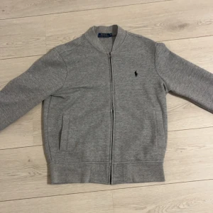 Grå tröja från Ralph Lauren - Säljer en stilren grå tröja från Ralph Lauren med dragkedja och ikonisk logga på bröstet. Perfekt för en avslappnad men snygg look. Tröjan har långa ärmar och är tillverkad i mjukt material för extra komfort.