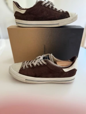 Bruna låga sneakers i mocka - Snygga bruna sneakers i mocka med vita detaljer och tåhätta. Skorna har vita snören och en klassisk låg siluett. Yttersulan är vit med en tunn rand i grått. Perfekta för dig som gillar stilrena och tidlösa sneakers.