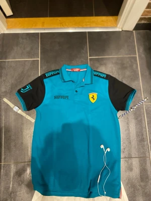 Blå Ferrari pikétröja special edition Juventus  - Snygg blå pikétröja från Ferrari med svarta ärmar och detaljer. Tröjan har klassisk krage, knappar framtill och Ferrari-logga på bröstet. Märkesnamnet syns på både bröst och axlar. Perfekt för dig som gillar sportig stil.