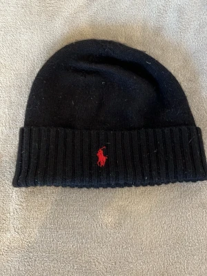 Svart mössa med röd logga - Snygg svart mössa från Polo Ralph Lauren med uppvikt kant och klassisk röd broderad logga framtill. Tillverkad i mjukt stickat material som håller dig varm och stilren under kalla dagar.