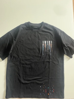 Svart AMIRI t-shirt - Svart t-shirt från AMIRI med korta ärmar och färgstänk i flera färger. Stort AMIRI-tryck på ryggen och mindre logga framtill, båda med droppande färgeffekt. Tillverkad i mjuk bomull och har en avslappnad passform.