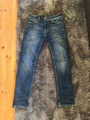 Blå tapered jeans från Jack & Jones - Snygga blå jeans från Jack & Jones i modellen Tapered Mike. Jeansen har en klassisk femficksdesign, smal passform och slitna detaljer för en trendig look. Tillverkade i denim med kontrastsömmar och dragkedjegylf.