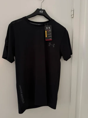 Svart träningströja från Under Armour - Helt ny med prislapp. En snygg svart t-shirt från Under Armour i storlek M med diskret logga på bröstet och tryck längs sidan. Tillverkad i ett lätt och funktionellt material som passar perfekt för träning. Klassisk rund hals och korta ärmar för en sportig look.