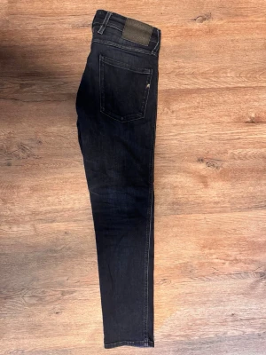 Mörkblå slim fit jeans från Replay - Snygga mörkblå slim fit jeans från Replay med modellen anbass och klassisk femficksdesign och subtila slitningar. Jeansen har en rak passform, knappgylf och detaljerade sömmar. Perfekta för en trendig och avslappnad look.