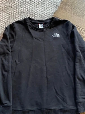 Svart sweatshirt från The North Face - Klassisk svart sweatshirt från The North Face med rund halsringning och logga på bröstet. Tröjan har långa ärmar och ribbade muddar vid ärmslut och nederkant. Storlek S