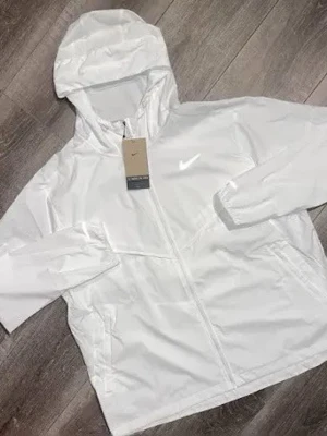 Vit vindjacka med huva från Nike - Snygg vit vindjacka från Nike med dragkedja framtill och praktisk huva. Jackan har två sidofickor och en minimalistisk design med Nike-logga på bröstet. Perfekt för blåsiga dagar och enkel att matcha till olika outfits.