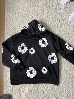 Denim tears - Säljer en svart hoodie med stora vita blommönster över hela tröjan och huvan. Hoodien har en klassisk känguruficka framtill och ribbade muddar vid ärmslut och nederkant. Perfekt för dig som gillar streetwear och vill sticka ut lite extra.