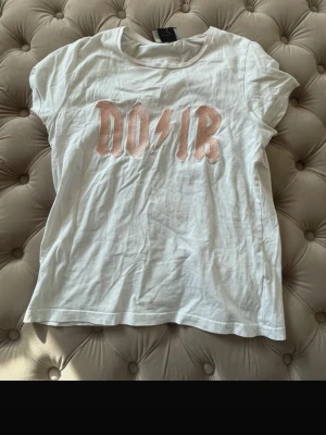 Vit t-shirt från Neo Noir - Vit t-shirt från Neo Noir med korta ärmar och trycket 'NOIR' i rosa med blixtdetalj på bröstet. Tillverkad i mjuk bomull och har en klassisk rund halsringning. Perfekt för en avslappnad och trendig look.