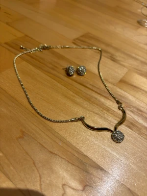 Guldigt halsband med pärldetalj - Säljer ett elegant halsband i guldfärgad metall med en böjd form och en rund hängande detalj täckt av små glittrande pärlor. Kedjan är tunn och smidig, och matchande örhängen med samma pärldesign ingår. Perfekt för dig som gillar stilrena accessoarer med lite extra bling.