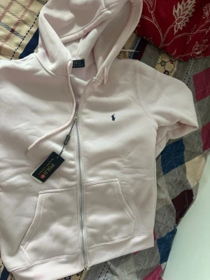 Ljusrosa hoodie från Polo Ralph Lauren - Ljusrosa hoodie från Polo Ralph Lauren med dragkedja, huva och kängurufickor. Klassisk broderad logga i mörkblått på bröstet. Mjuk bomullsblandning som känns skön mot huden. Perfekt för en chill och stilren look.
