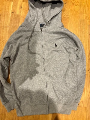Grå zip hoodie från Polo Ralph Lauren - Snygg grå hoodie med dragkedja från Polo Ralph Lauren. Klassisk design med huva och justerbara snören, samt den ikoniska loggan broderad på bröstet. Tillverkad i mjukt bomullsmaterial och har ribbade muddar vid ärmar och nederkant.