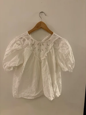 Vit blus med spetsdetaljer - Söt vit blus med puffiga korta ärmar och rund halsringning. Framtill finns en bred spetskant med broderade detaljer som ger en romantisk känsla. Tillverkad i ett lätt tyg, perfekt för en trendig och avslappnad look.